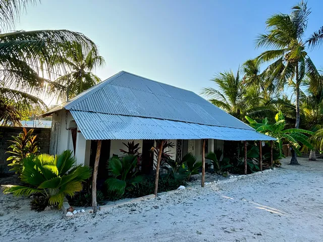 The house from outside: blue roof among palm trees, sandy path in front